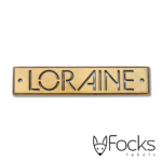 Loraine merklabel, gegoten zinklegering waarbij logo is uitgespaard, in antiek messing look vernikkeld, 2 boorgaten voor bevestiging.